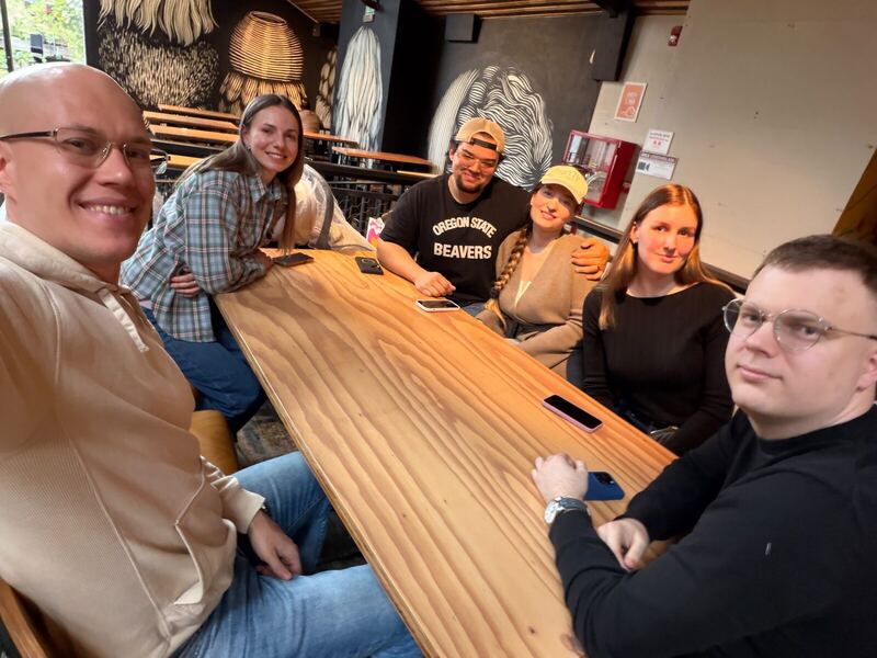 Six community members smiling around a long wooden table in a cafe